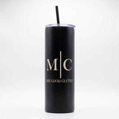 A matte black insulated tumbler with a clear lid and black straw, featuring the gold printed initials 'M|C' and the text 'THE GOOD GLUTEN' on the front, set against a plain white background.