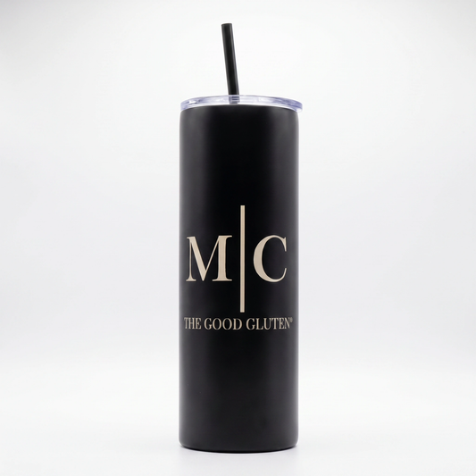A matte black insulated tumbler with a clear lid and black straw, featuring the gold printed initials 'M|C' and the text 'THE GOOD GLUTEN' on the front, set against a plain white background.